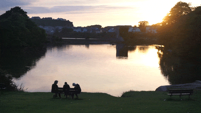 Three people sit by a lake at sunset, one of them is eating something